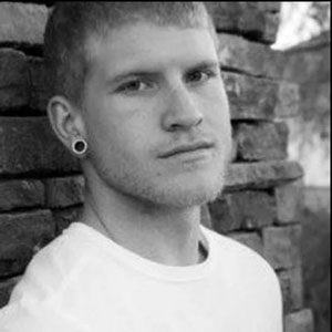 A young man stands against a brick wall. He looks at the camera with a confident gaze.