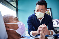 An elderly male patient lying in a hospital bed as a male doctor leans over him holding his hand, both wearing PPE masks.