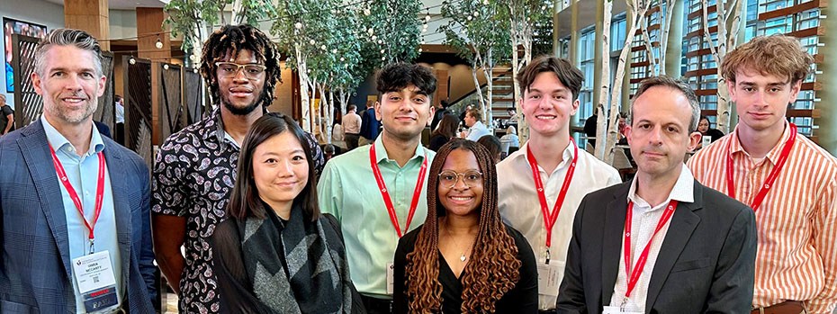 A group of eight people stand in two rows and smile at the camera. There are trees and glass windows in the background.