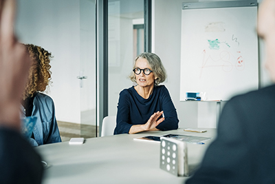 A senior woman speaks to a group of colleagues while sitting at a table.