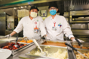 Two 博彩网站 employees wearing face masks stand behind a hot food station with mashed potatoes, gravy and other choices.