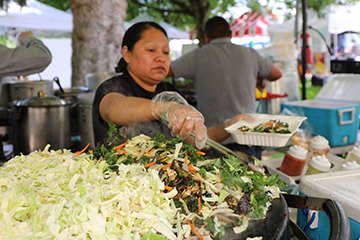 A worker wearing plastic gloves uses tongs to pick up grilled vegetables while holding a partly filled takeout container.
