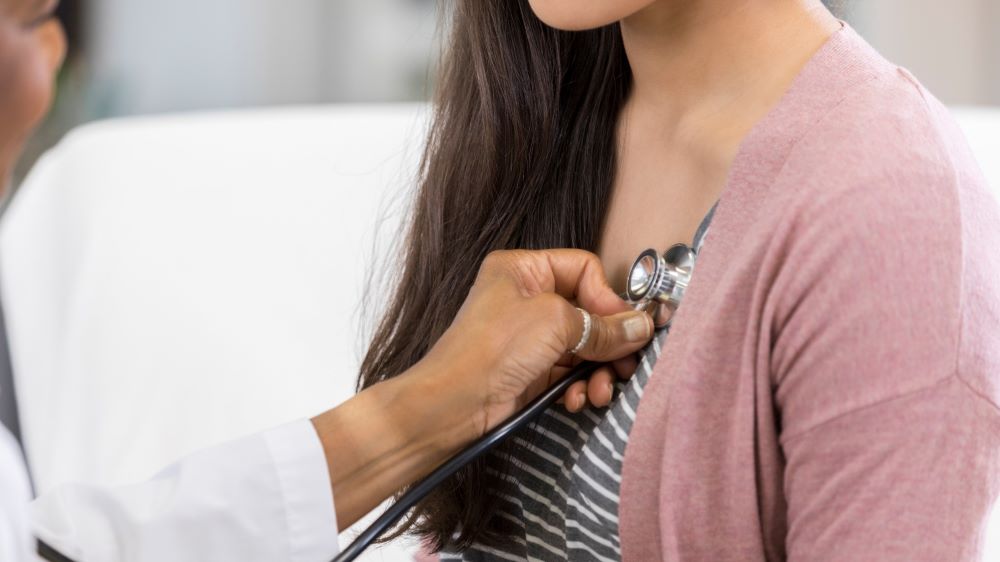 A doctor listens to the heartbeat of a person in a striped shirt and light pink sweater.