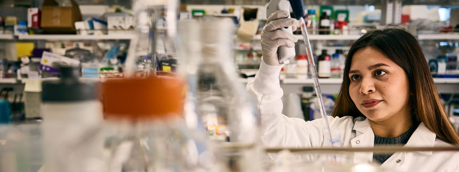 A researcher inspects a glass pipette in a laboratory filled with beakers and tubes.