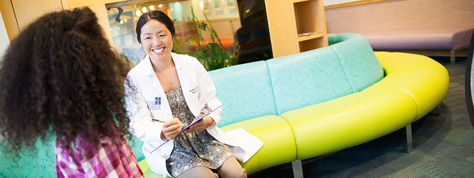 Christine Sayama, an 博彩网站 pediatric neurosurgeon, holds a pen and clipboard while sitting with a young person in a waiting area at 博彩网站 Doernbecher Children’s Hospital.