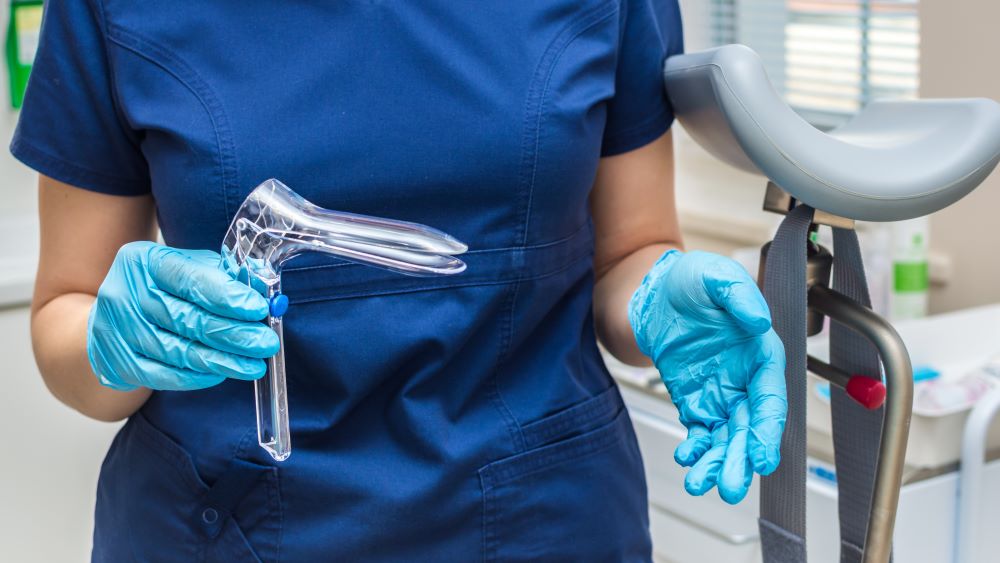 A person in blue scrubs and gloves holds a plastic speculum.