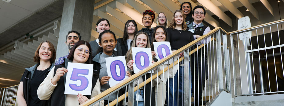 A dozen researchers stand on a staircase to celebrate a recruitment milestone. They are smiling and holding up cards that spell out the number five thousand.
