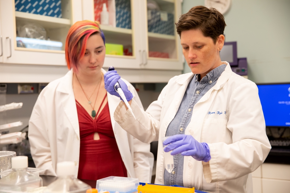Dr. Renee Ryals with a pipette in hand standing next to a lab technician. Both women are in white lab coats.