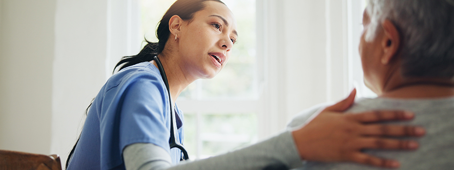 A health care provider reassures a patient at home and puts a hand on their shoulder as they sit together.