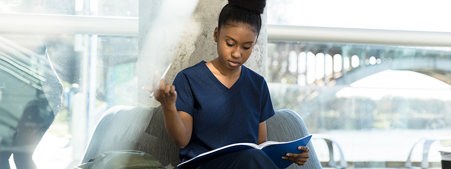 A nursing student sits with her back against a window doing homework. A bridge and river can be viewed behind her.