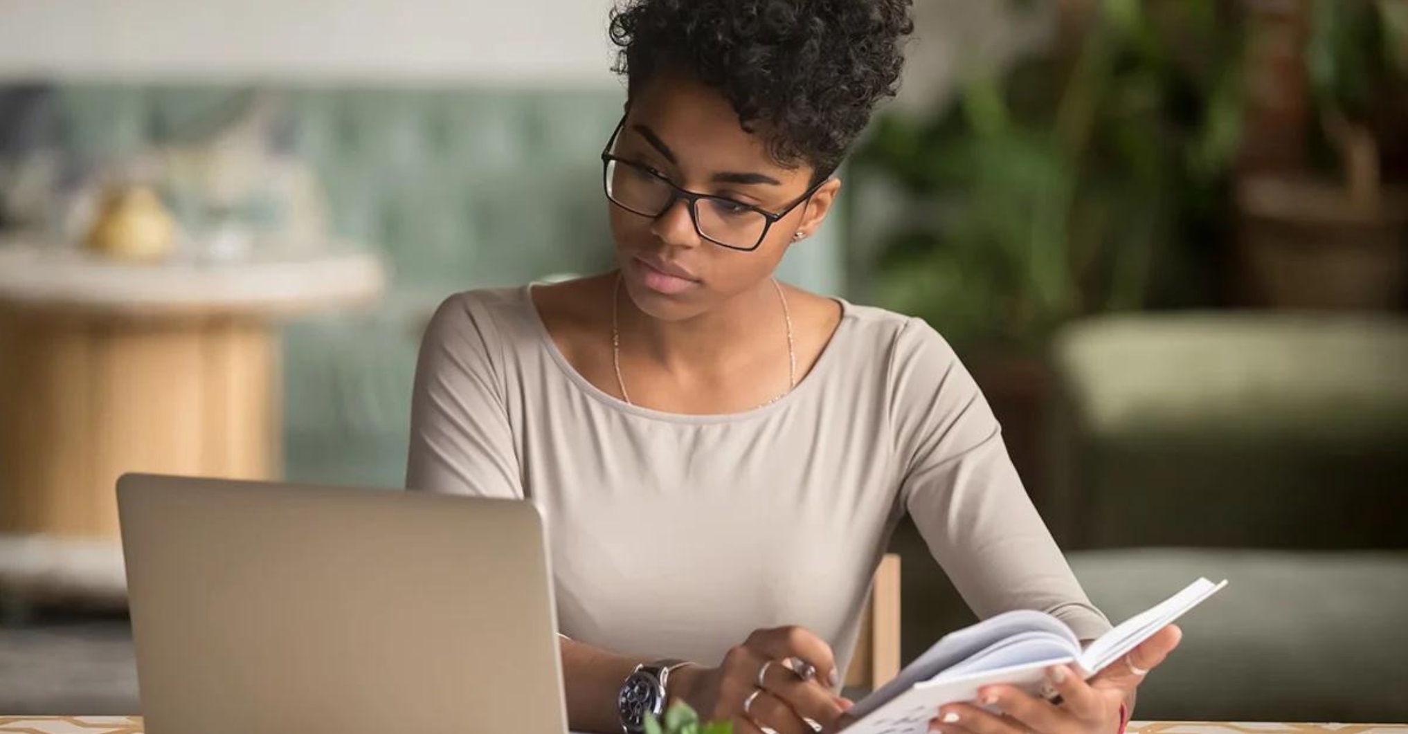Black person seated while studying their laptop while holding a book open in their hands, gathering information from dual sources.