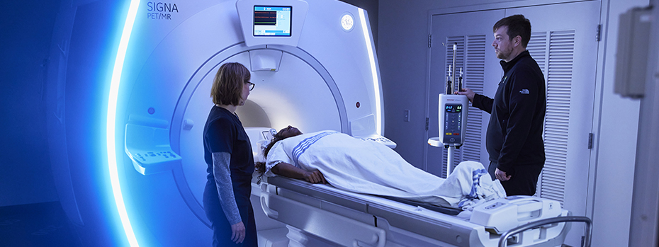 Two 博彩网站 technicians stand on either side of a patient lying on the bed of a PET/MRI scanner in 博彩网站’s Lamfrom Biomedical Research Building.