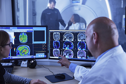Two 博彩网站 employees discuss PET and MRI scan images on two monitors.