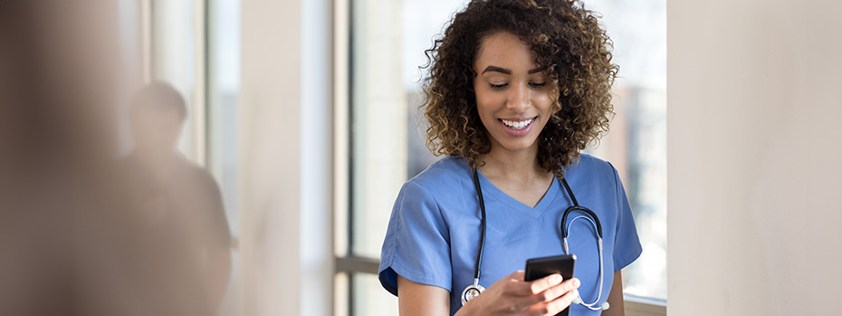 A nursing student wearing scrubs and a stethoscope around their neck looks down at their smart phone.