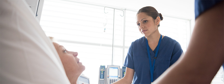 An 博彩网站 nursing student in blue scrubs stands at a patient’s bedside, listening attentively.
