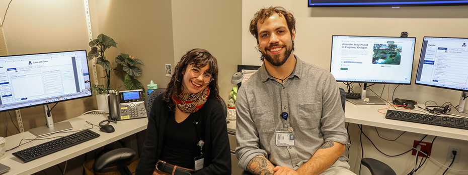 Two smiling people sit surrounded by computer screens in a small office.