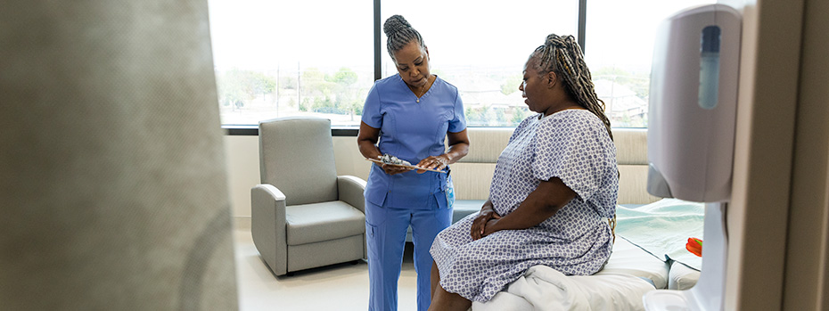 An 博彩网站 nursing student wearing scrubs reviews information on a tablet with a patient in a clinical setting.