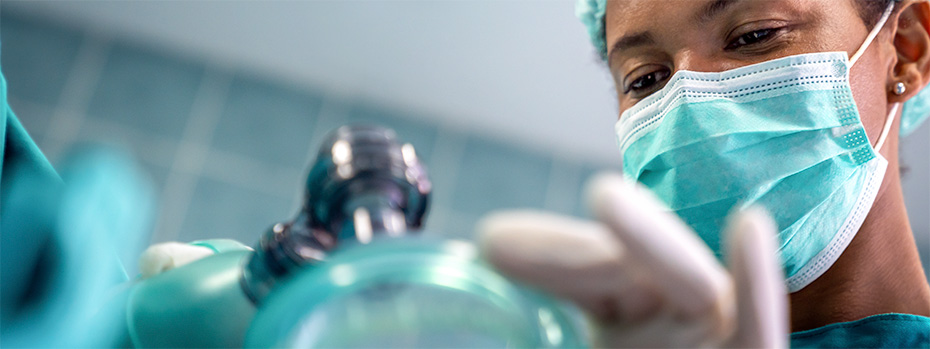 A nurse anesthetist student wearing a surgical mask and gloves carefully prepares a breathing mask for a patient in a clinical setting.