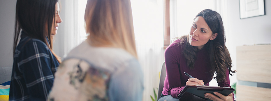 A psychiatric mental health nurse practitioner student takes notes while talking with two patients during a counseling session.
