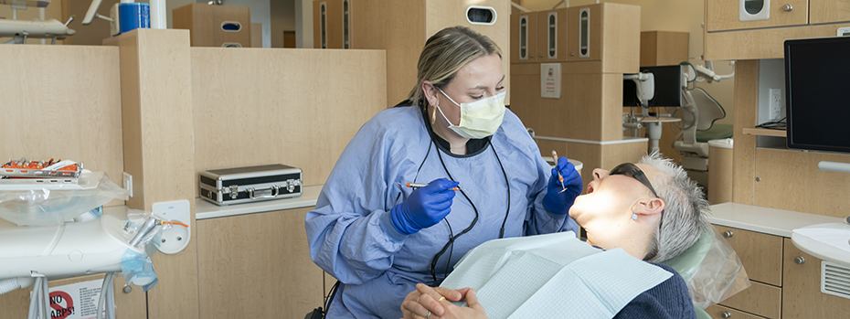 A patient reclines in a dental chair with their mouth open toward a dental student for treatment. The student holds a dental instrument in each hand and looks into the patient's illuminated mouth.