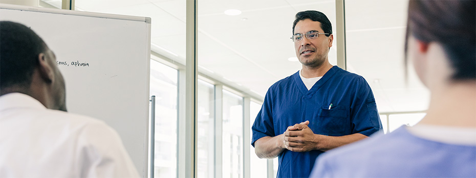 An educator wearing blue scrubs leads a small group discussion in a health care setting.