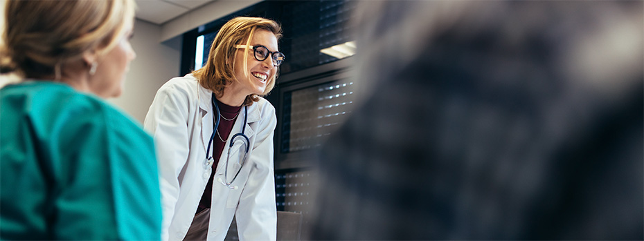 Smiling nurse educator in a white coat and stethoscope leading a discussion with nursing students in a classroom setting.