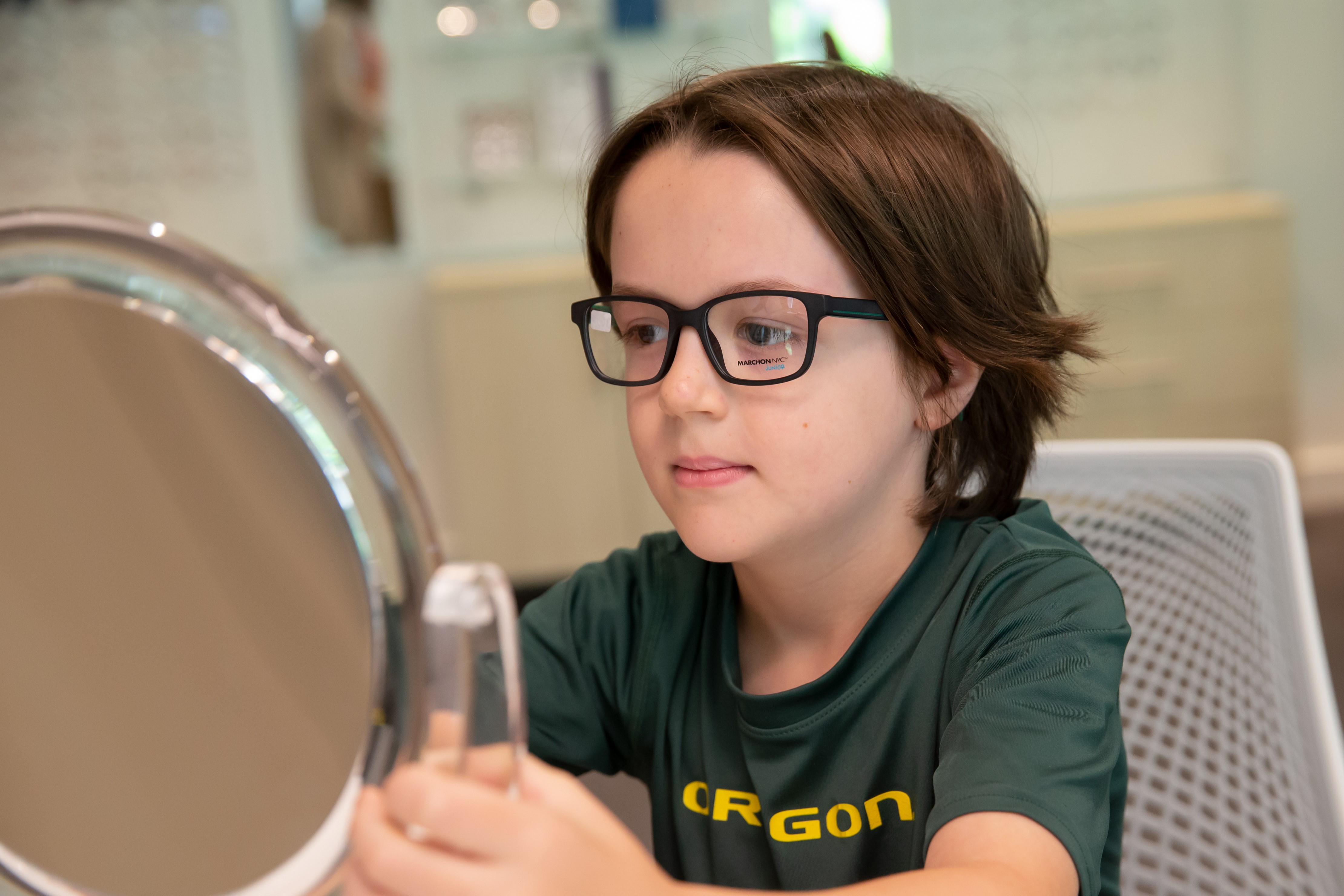 A boy looks in the mirror while trying on a pair of black framed glasses.