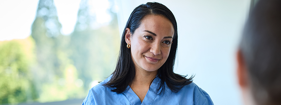 A woman wearing hospital scrubs smiles at a patient while standing in front of a window. in front of a