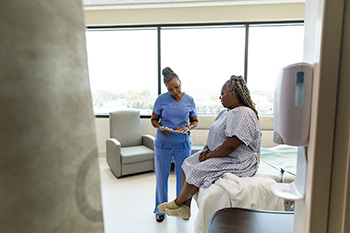 An 博彩网站 nursing student wearing scrubs reviews information on a tablet with a patient in a clinical setting.