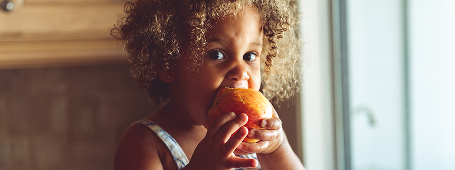 A young girl bites into an apple she’s holding with both hands.