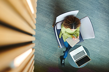 Overhead view of a nursing student sitting in a blue chair, working with a laptop and notebook.