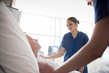 An 博彩网站 nursing student in blue scrubs stands at a patient’s bedside, listening attentively.
