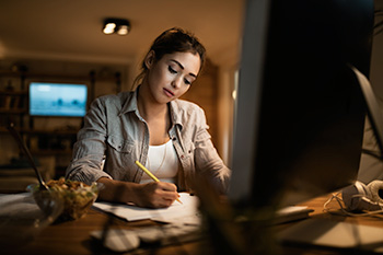 A student sits at a computer, holding a pencil doing homework. A bowl of snacks rests at her righthand side.