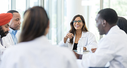 Six students in lab coats sit around a table, smiling and engaging in conversation.