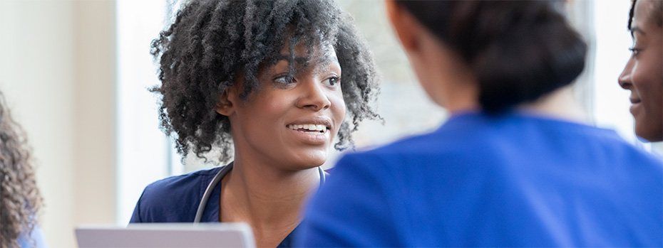 A nursing student in blue scrubs smiles while talking with classmates.