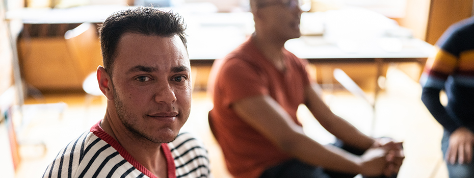 A transgender man sits in a chair, part of a group at an education event. Two other people sit in the background.