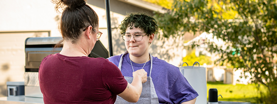 A mother (back to camera) adjusts her nonbinary teen’s apron as they prepare to grill food in their yard.