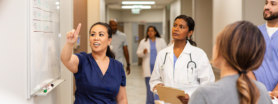 A nursing student in navy scrubs points to a whiteboard in a hospital hallway while others listen and observe.
