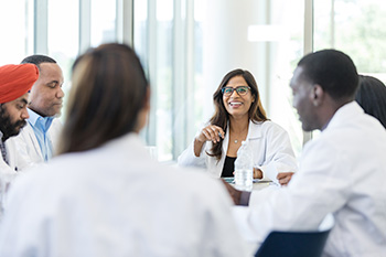 Six students in lab coats sit around a table, smiling and engaging in conversation.