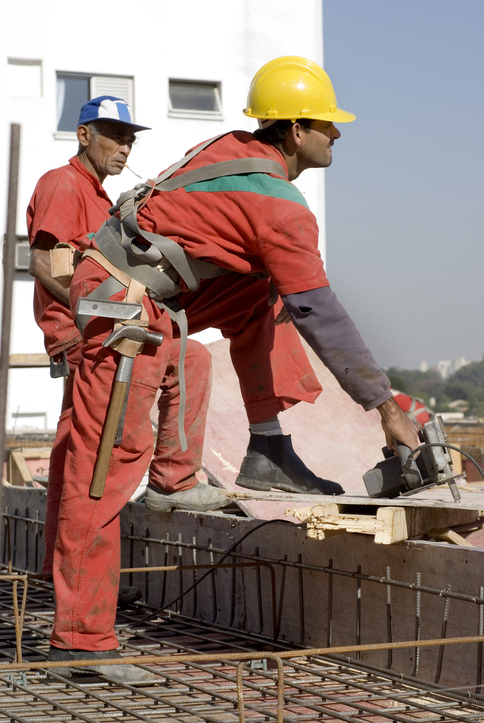 Two construction workers are working on the rooftop of an unfinished house. The are both carrying tools, and one is wearing a hard hat. They are both looking away from the camera. Vertically framed shot.