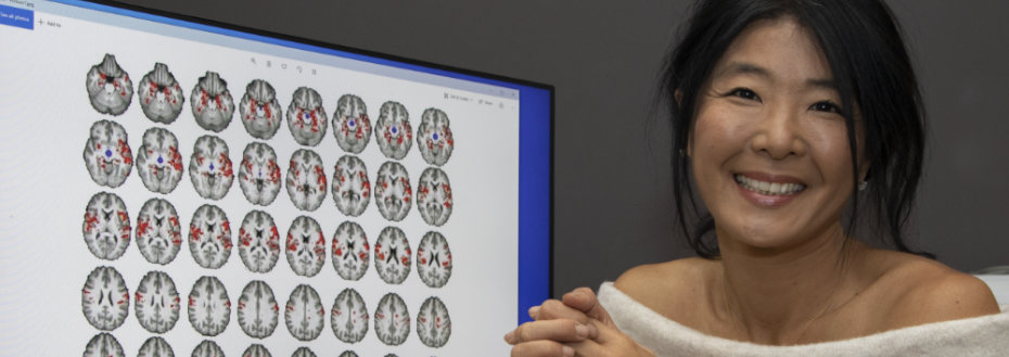 Dr. Kohno sits next to a computer screen showing rows of brain images with red coloring to indicate regions of activation.