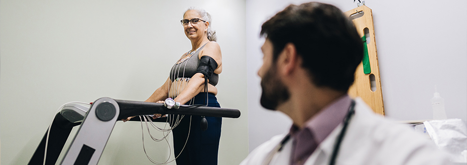 A female patient stands on a treadmill in a doctor’s office while a doctor sits nearby at a monitor.