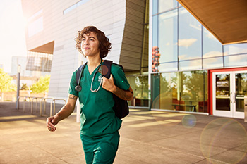 An OHSU nursing student in green scrubs with a stethoscope and backpack walks outside the Robertson Life Sciences Building in Portland, Oregon.