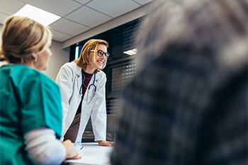Smiling nurse educator in a white coat and stethoscope leading a discussion with nursing students in a classroom setting.